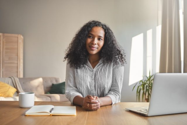 portrait-positive-self-confident-young-dark-skinned-female-teacher-with-voluminous-hairdo-getting-ready-online-lesson-sitting-desk-with-laptop-coffee-copybook-home-office-interio